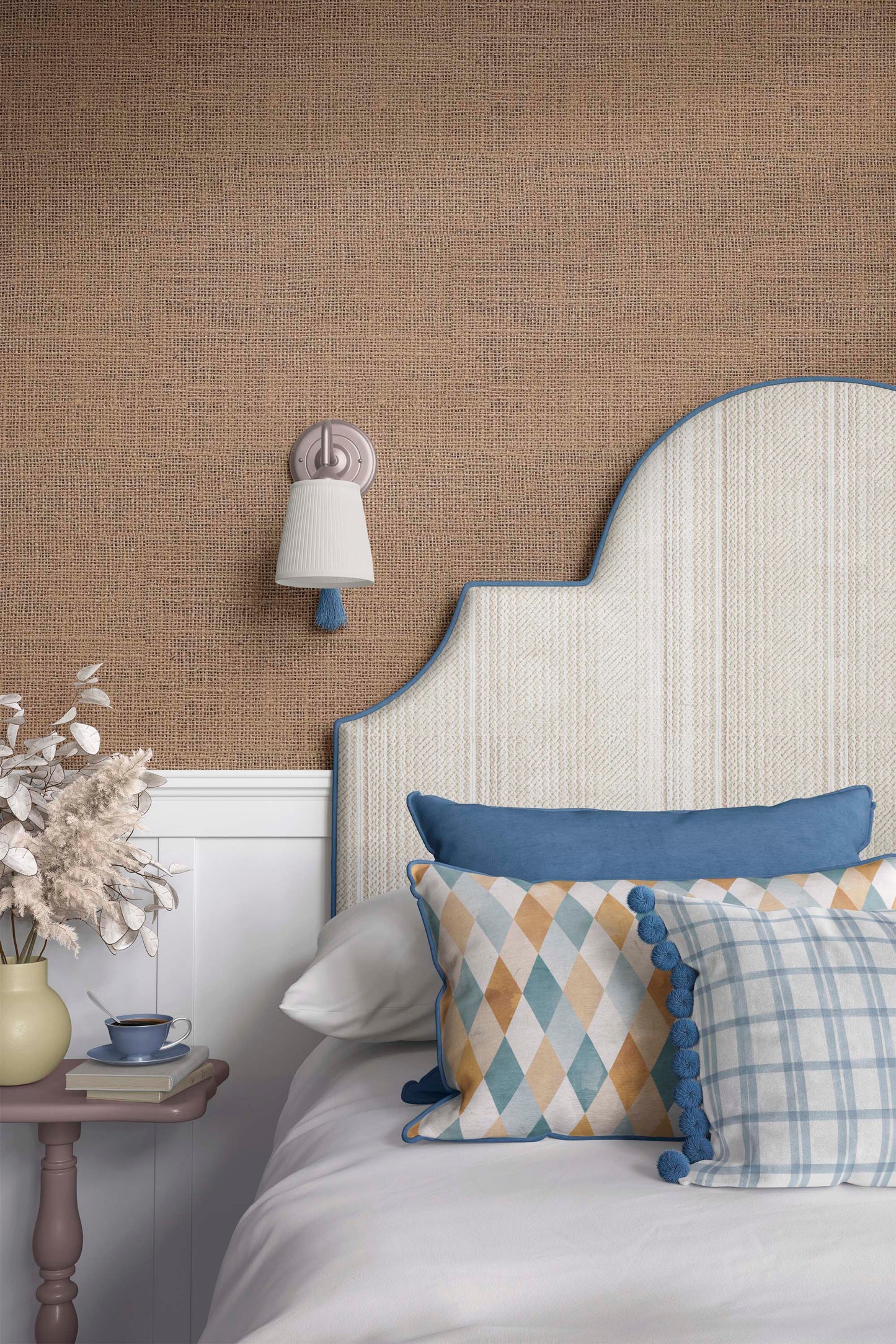 A cozy bedroom setup featuring a decorative headboard with a blue trim, patterned pillows on a white bed, a bedside table with a small cup and decorative flowers, and a soft light fixture on the wall against a textured brown background.