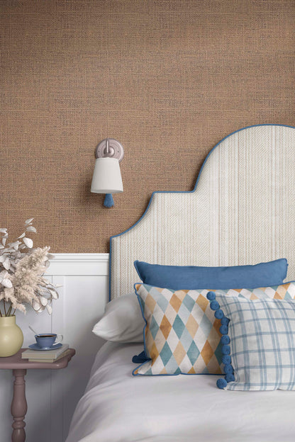 A cozy bedroom setup featuring a decorative headboard with a blue trim, patterned pillows on a white bed, a bedside table with a small cup and decorative flowers, and a soft light fixture on the wall against a textured brown background.