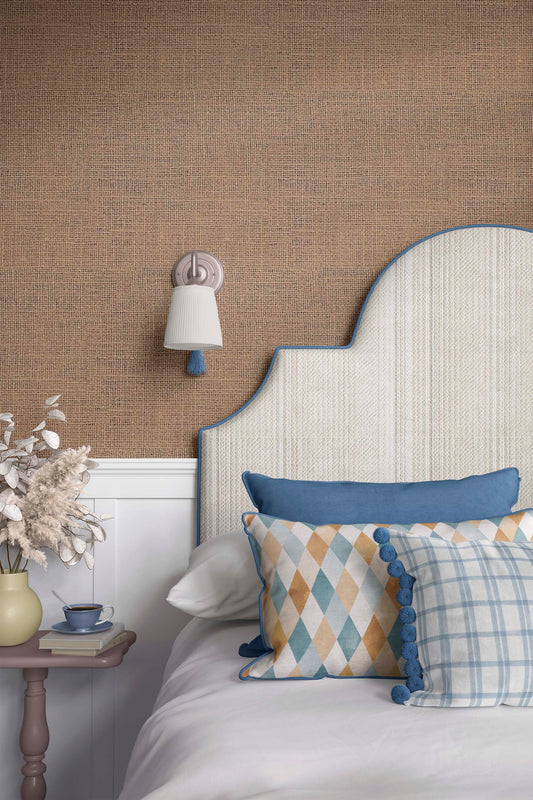 A cozy bedroom setup featuring a decorative headboard with a blue trim, patterned pillows on a white bed, a bedside table with a small cup and decorative flowers, and a soft light fixture on the wall against a textured brown background.