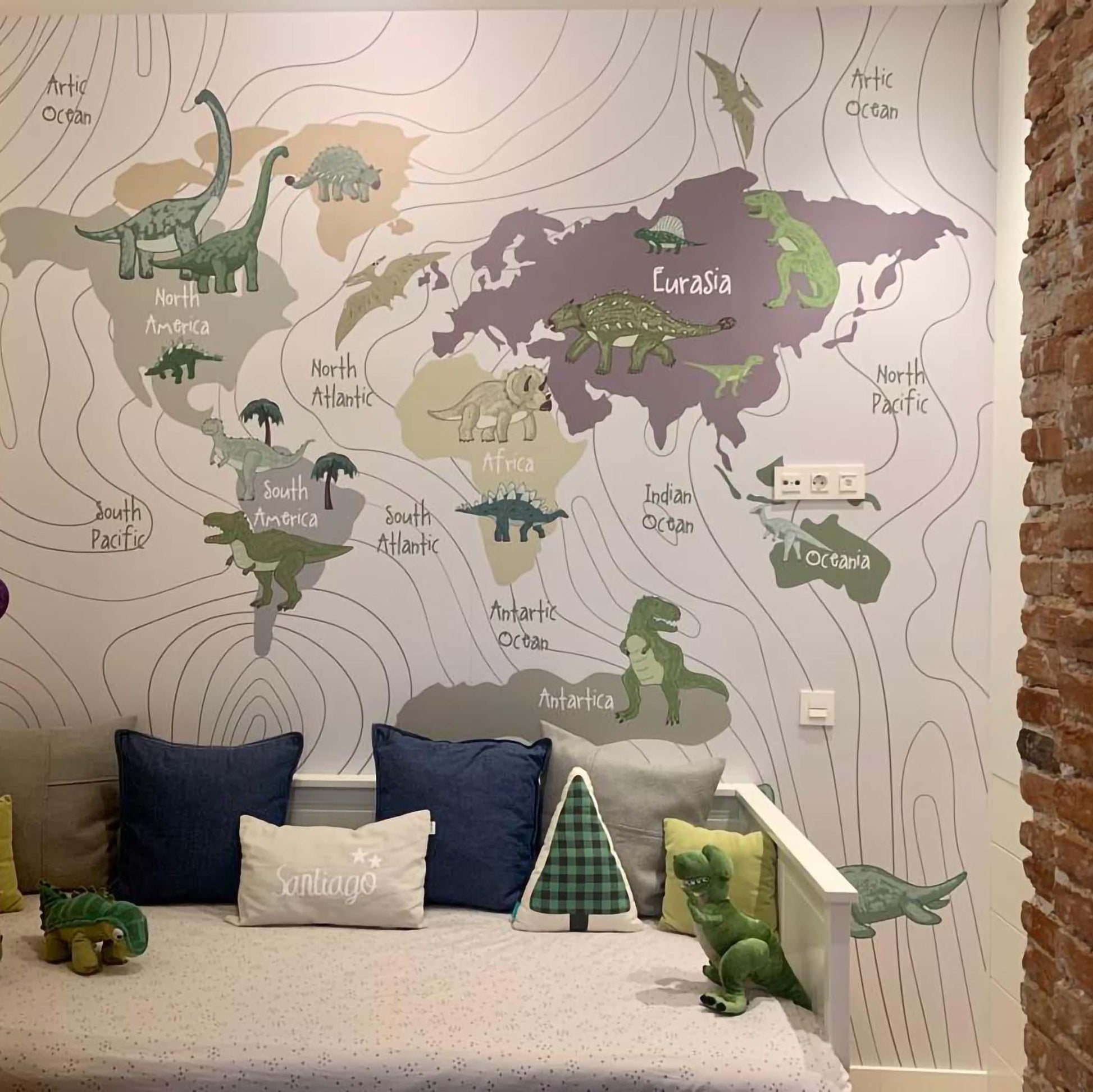 A cozy children's nook framed by brick walls featuring a world map dinosaur mural, a white daybed with navy and grey pillows, and colorful string lights.