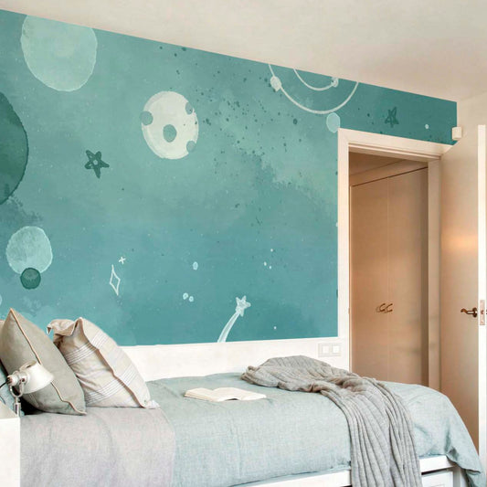 A peaceful bedroom featuring a large teal watercolor space mural with white stars and moons, a bed with light blue bedding, and a cozy grey knit blanket.
