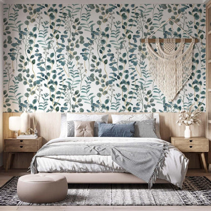 cat-616983200072 A bright bedroom featuring a teal and white botanical wallpaper with watercolor eucalyptus leaves as a central accent wall. A large cream macramé wall hanging is centered above a bed with neutral-toned bedding and a patterned throw. The room is completed with light wood furniture, a textured rug, and a minimalist aesthetic.