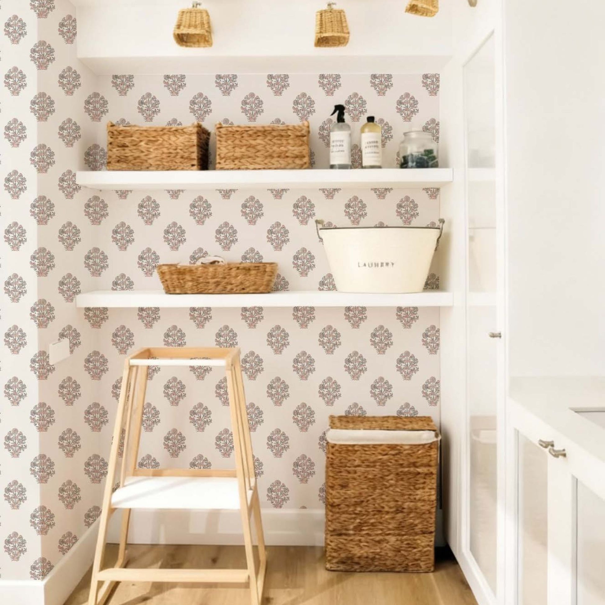 cat-615198490952 An organized and airy laundry room featuring a floral block print wallpaper with pink and charcoal buta motifs. The decorative accent wall is fitted with white floating shelves holding several woven seagrass baskets, a vintage-style white metal laundry bucket, and glass cleaning jars. A wooden step stool and a matching tall wicker hamper sit on the light wood flooring, creating a harmonious and functional workspace