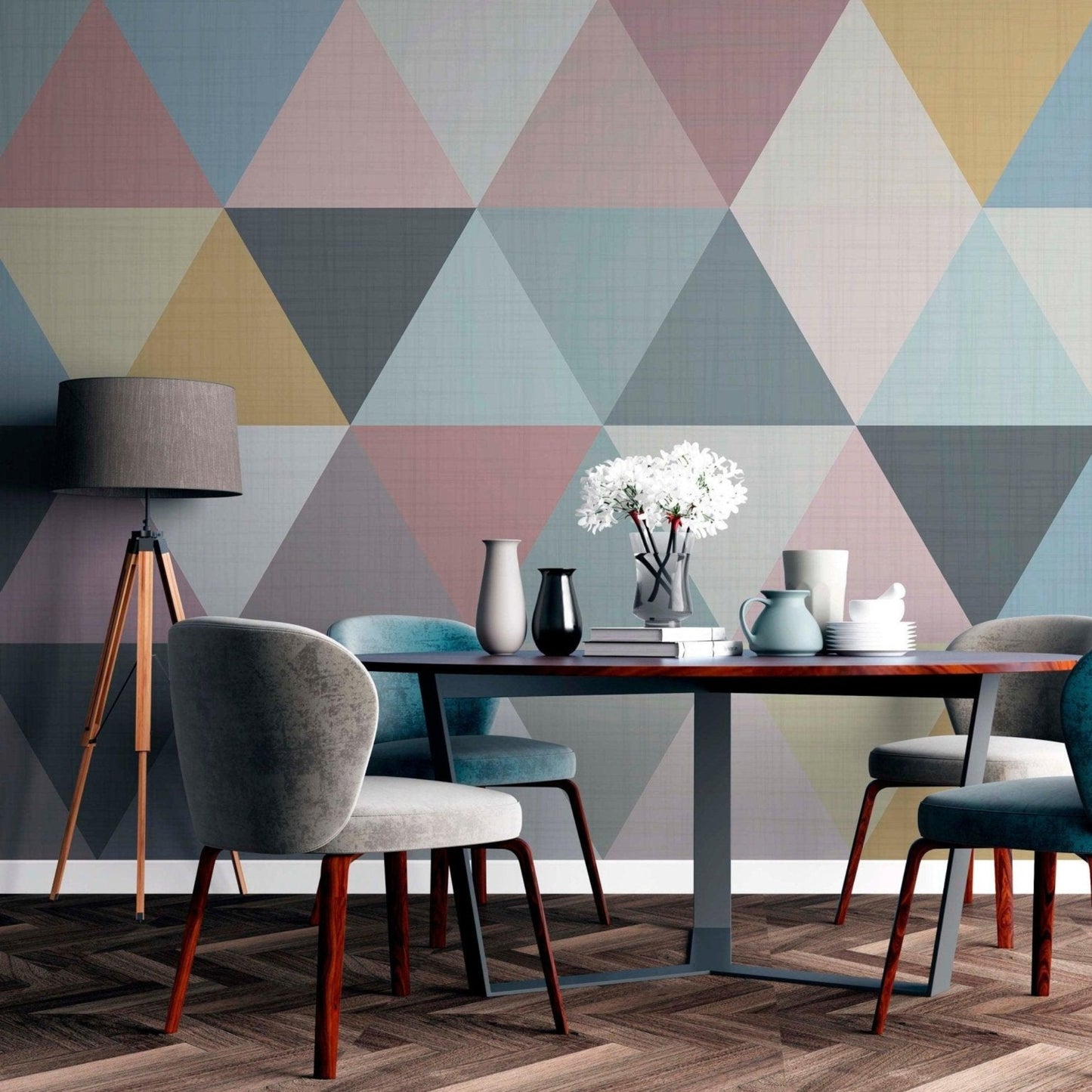cat-610512830792 A modern dining room featuring a pastel geometric triangle wallpaper mural behind a dark wood dining table with upholstered grey and blue chairs, a tripod floor lamp, and herringbone flooring.
