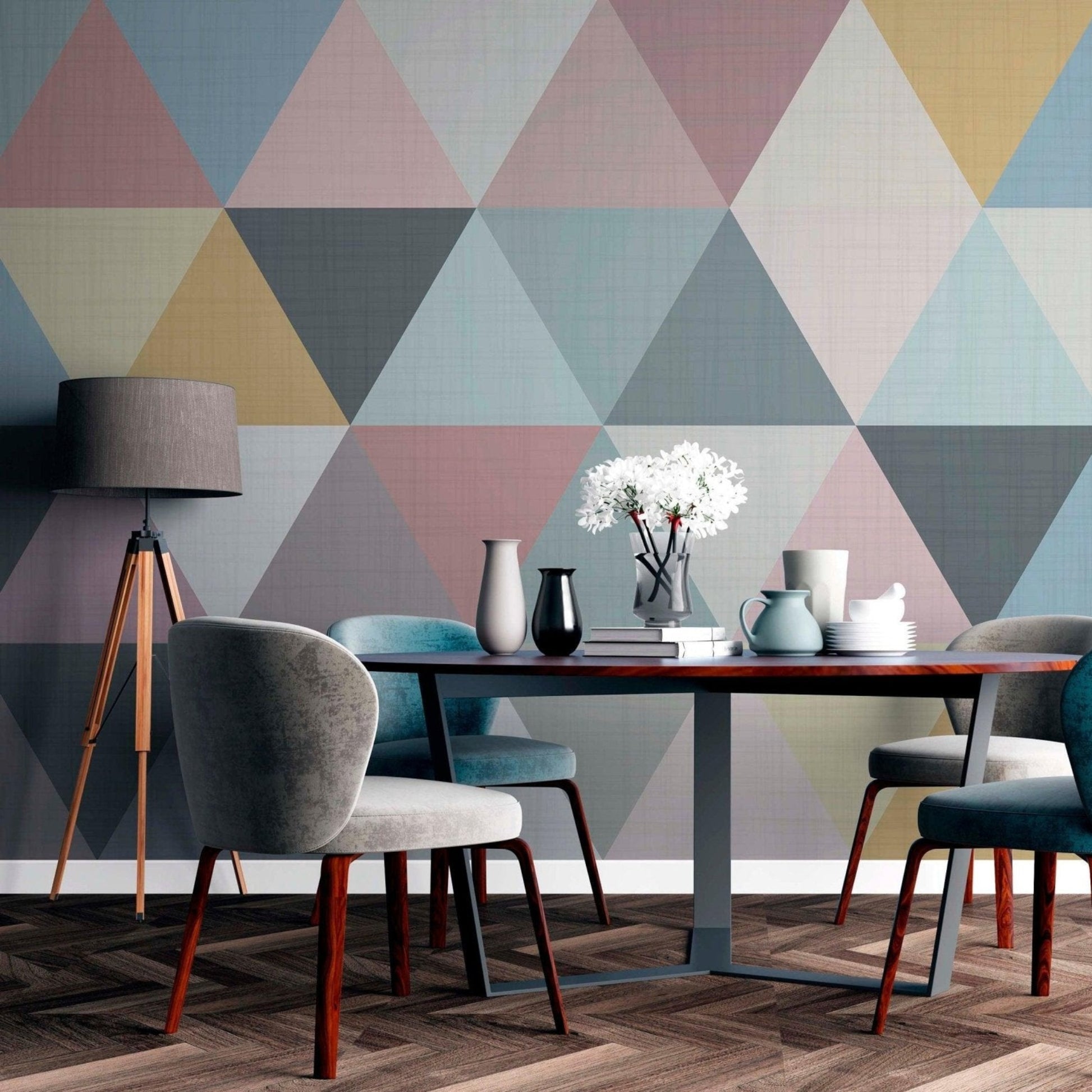 cat-610512830792 A modern dining room featuring a pastel geometric triangle wallpaper mural behind a dark wood dining table with upholstered grey and blue chairs, a tripod floor lamp, and herringbone flooring.