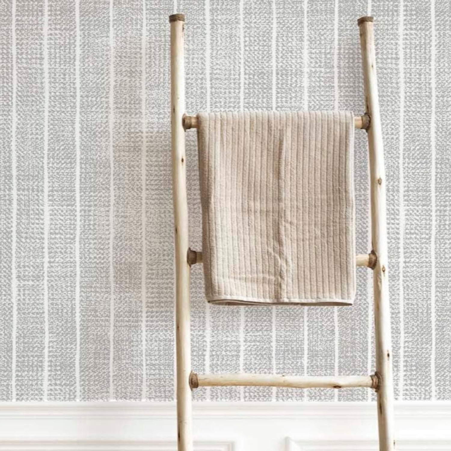 Bright laundry room featuring grey textured vertical stripe wallpaper above white wainscoting, with a white dryer, a wooden towel ladder, and a wicker laundry basket on herringbone floors