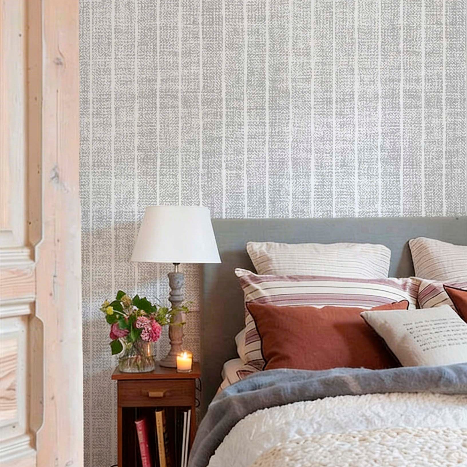 cat-616983200072 Cozy bedroom with grey and white woven-effect stripe wallpaper. The bed features earthy terracotta and striped pillows, a grey headboard, and a chunky knit throw, next to a wooden nightstand with a lamp and flowers