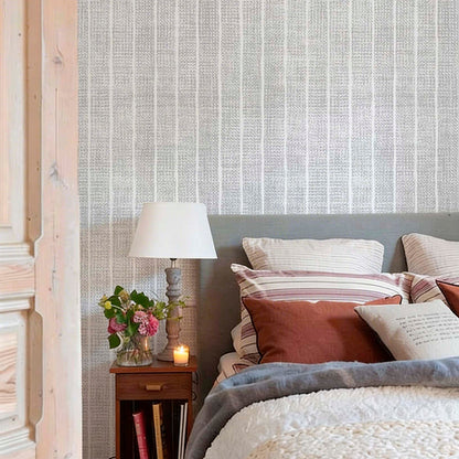 cat-616983200072 Cozy bedroom with grey and white woven-effect stripe wallpaper. The bed features earthy terracotta and striped pillows, a grey headboard, and a chunky knit throw, next to a wooden nightstand with a lamp and flowers