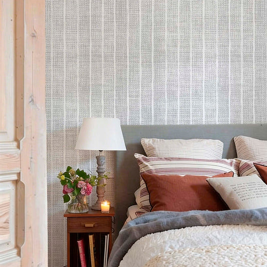 cat-616983200072 Cozy bedroom with grey and white woven-effect stripe wallpaper. The bed features earthy terracotta and striped pillows, a grey headboard, and a chunky knit throw, next to a wooden nightstand with a lamp and flowers