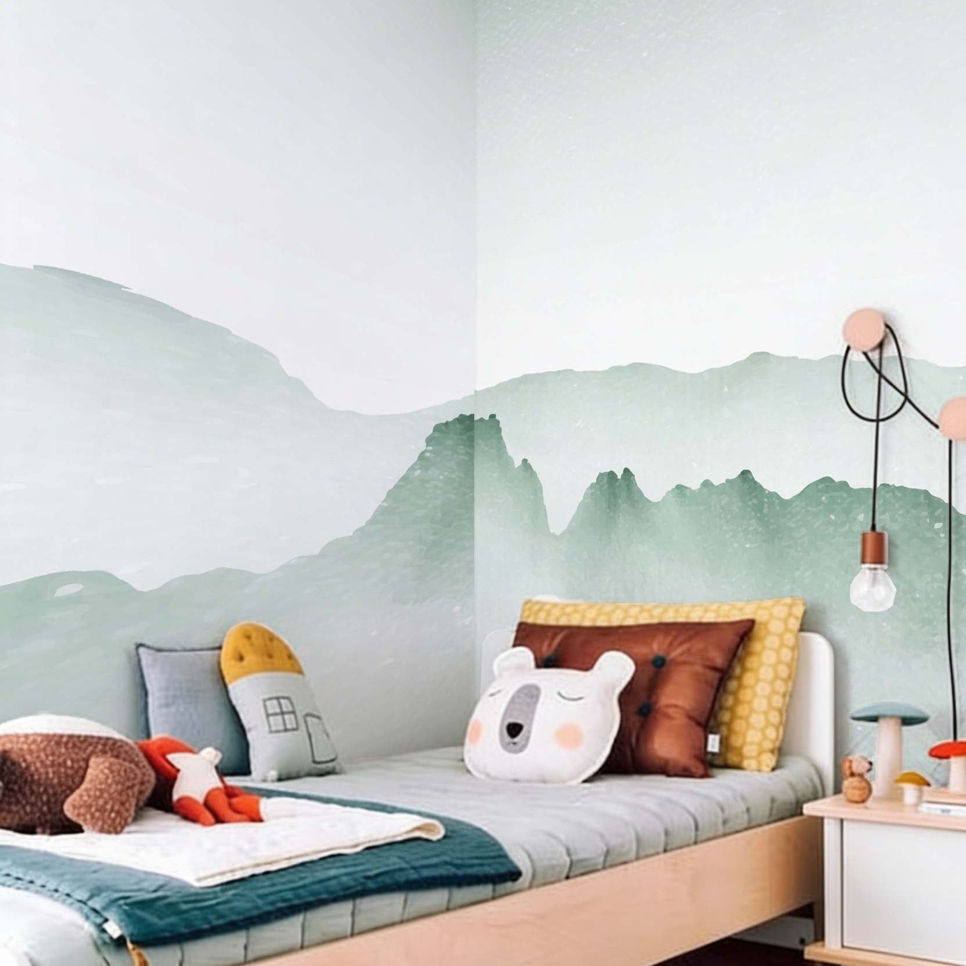 Decorative kids wallpaper featuring soft green watercolor mountains on a bedroom wall corner next to a light wood bed with mustard and animal cushions.