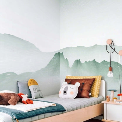 Decorative kids wallpaper featuring soft green watercolor mountains on a bedroom wall corner next to a light wood bed with mustard and animal cushions.