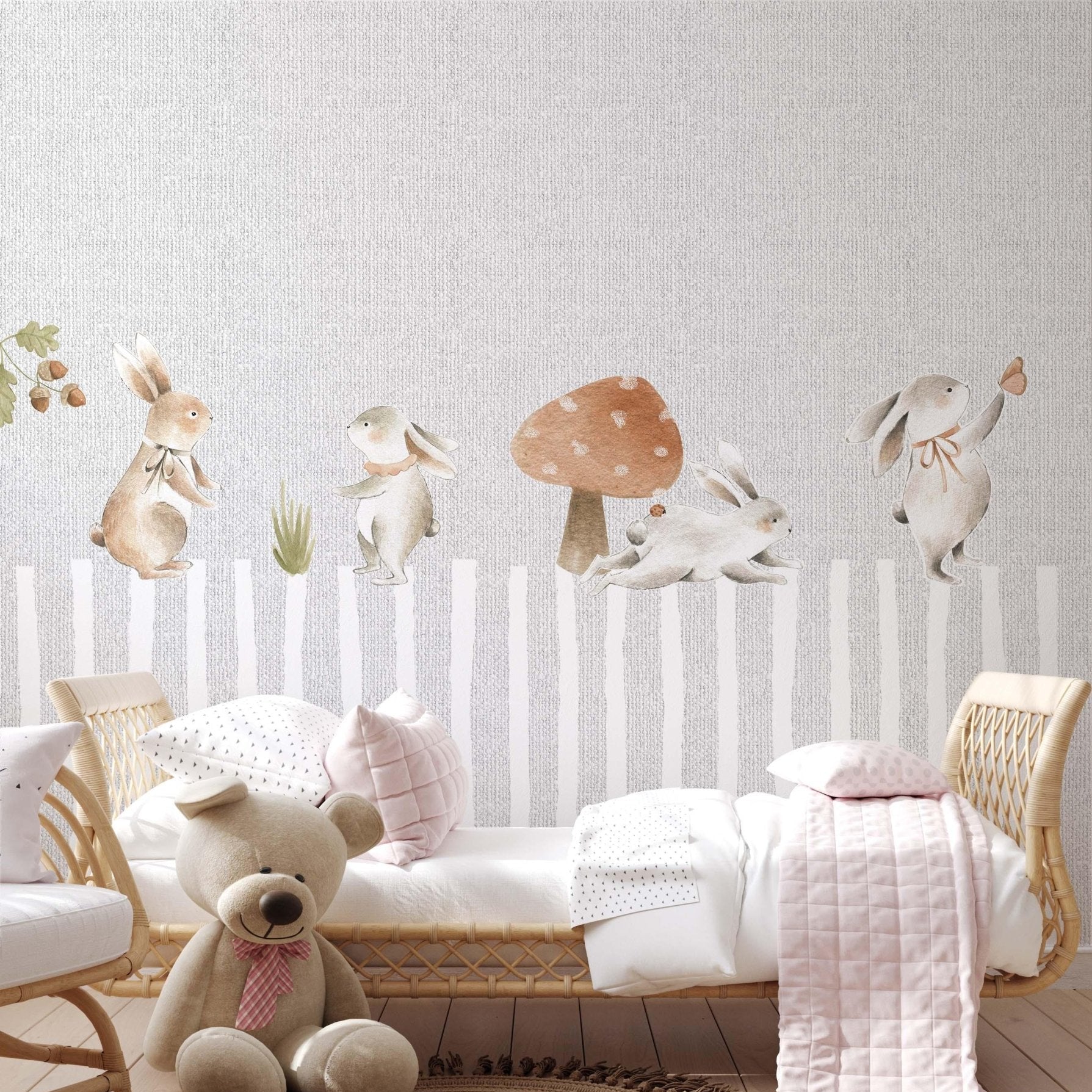 Decorative kids wallpaper featuring watercolor forest bunnies over a grey faux linen textured striped background on a bedroom wall behind a natural rattan toddler bed with a large teddy bear.