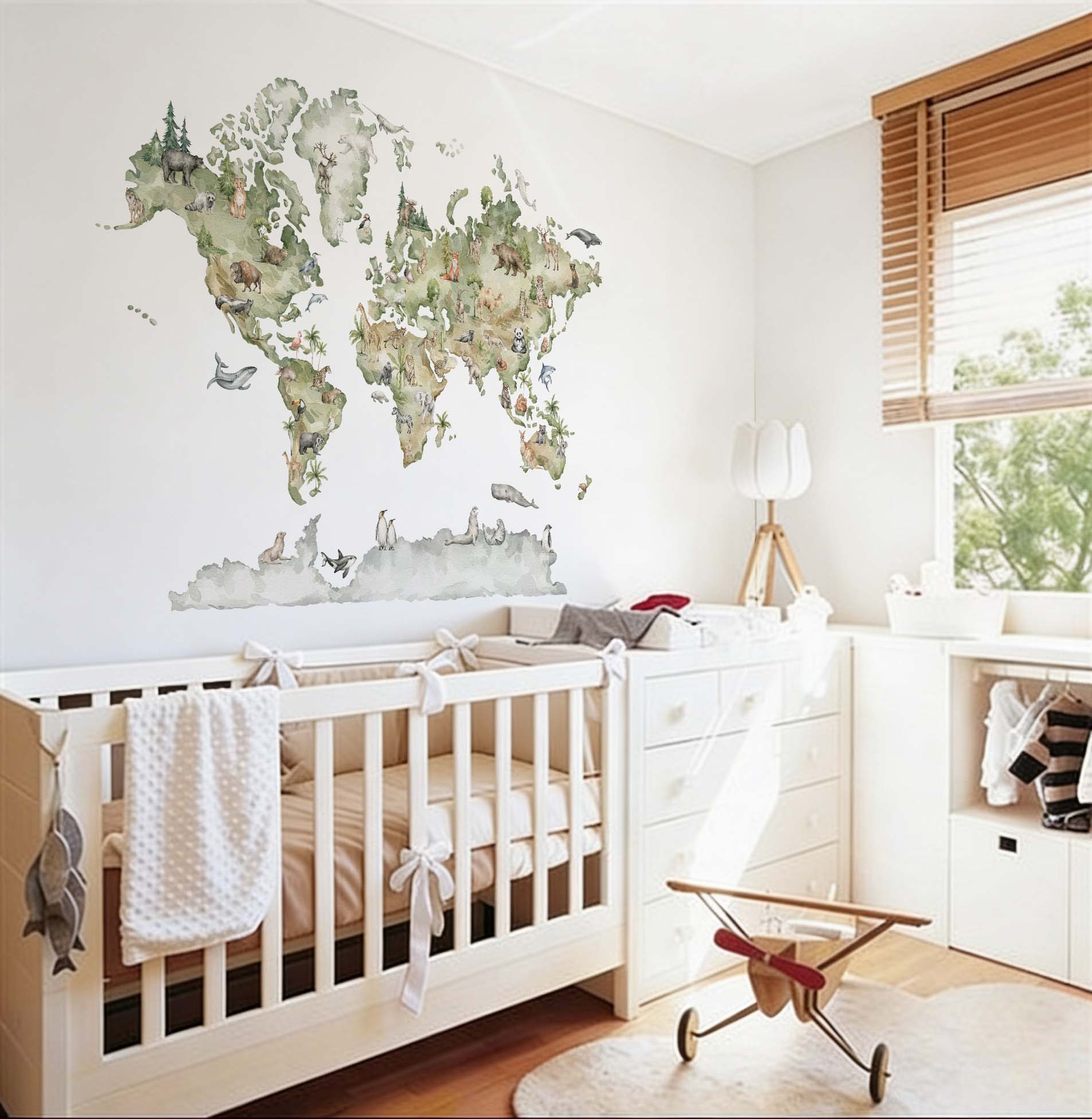 Watercolor animals world map wallpaper mural installed in a bright baby bedroom featuring a white wooden crib, dresser, and a vintage toy plane.