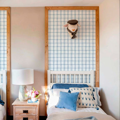 Close-up of light blue and white plaid patterned wallpaper used as a decorative wall inlay, framed with natural wood molding above a white wooden headboard.