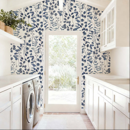 cat-615198490952 A bright and airy laundry room features a striking accent wall covered in navy blue botanical wallpaper. The pattern consists of delicate, watercolor-style eucalyptus leaves and leafy stems set against a clean white background, framing a central glass door that leads to a sunlit outdoor space. The deep blue tones of the wallpaper provide a bold contrast to the room’s crisp white cabinetry, light countertops, and neutral-colored flooring.