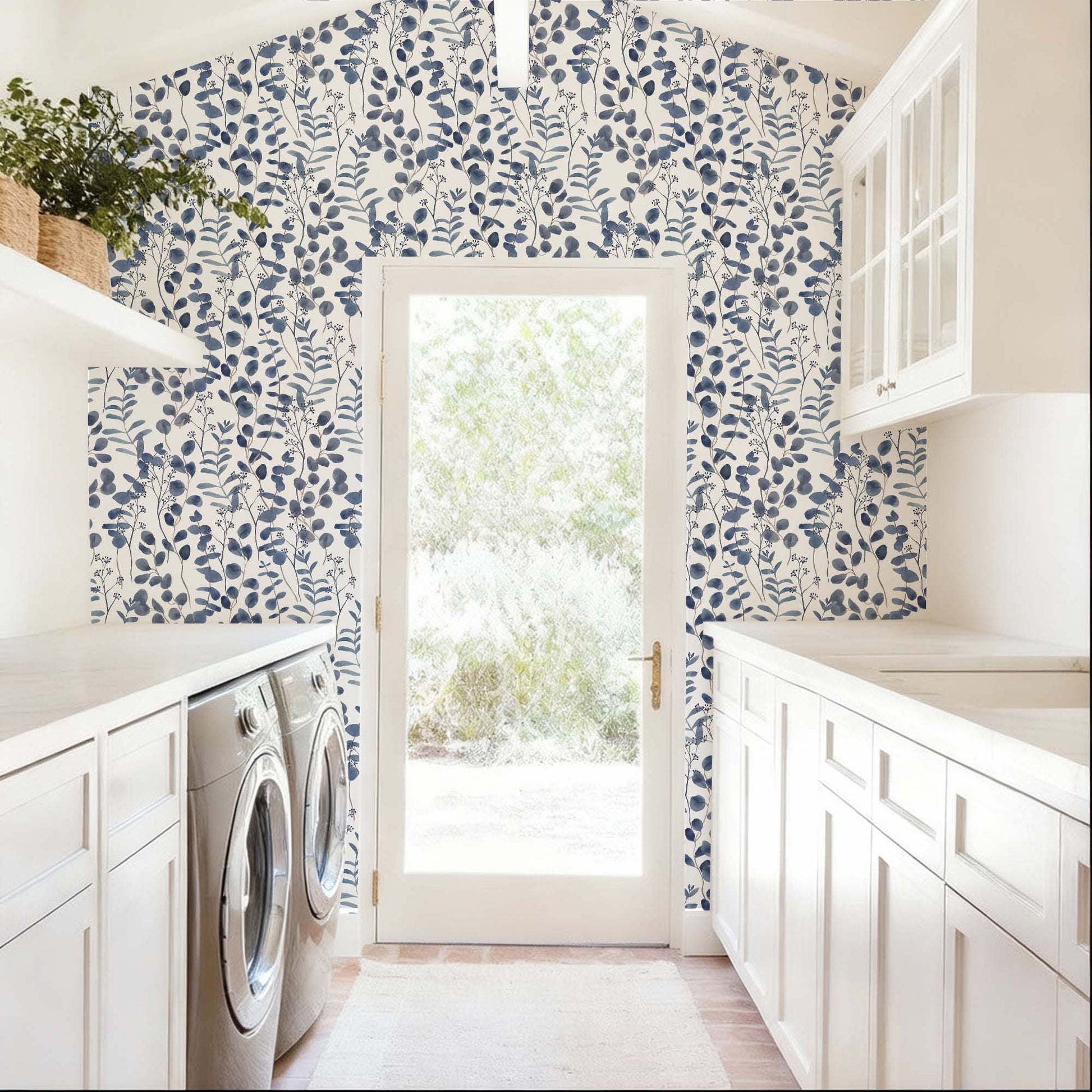 cat-615198490952 A bright and airy laundry room features a striking accent wall covered in navy blue botanical wallpaper. The pattern consists of delicate, watercolor-style eucalyptus leaves and leafy stems set against a clean white background, framing a central glass door that leads to a sunlit outdoor space. The deep blue tones of the wallpaper provide a bold contrast to the room’s crisp white cabinetry, light countertops, and neutral-colored flooring.