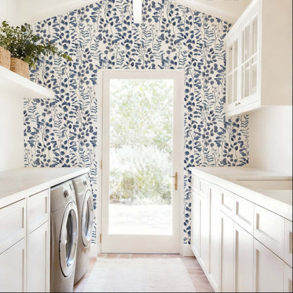 cat-615198490952 A bright and airy laundry room features a striking accent wall covered in navy blue botanical wallpaper. The pattern consists of delicate, watercolor-style eucalyptus leaves and leafy stems set against a clean white background, framing a central glass door that leads to a sunlit outdoor space. The deep blue tones of the wallpaper provide a bold contrast to the room’s crisp white cabinetry, light countertops, and neutral-colored flooring.