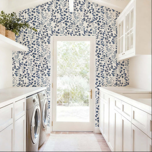 cat-615198490952 A bright and airy laundry room features a striking accent wall covered in navy blue botanical wallpaper. The pattern consists of delicate, watercolor-style eucalyptus leaves and leafy stems set against a clean white background, framing a central glass door that leads to a sunlit outdoor space. The deep blue tones of the wallpaper provide a bold contrast to the room’s crisp white cabinetry, light countertops, and neutral-colored flooring.