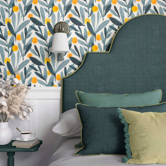 A vibrant botanical wallpaper featuring watercolor-style blue-green leaves and bright yellow citrus accents, creating a cheerful and fresh atmosphere. The wallpaper is styled in a cozy bedroom with a teal upholstered headboard and matching pillows, complemented by a modern wall sconce.