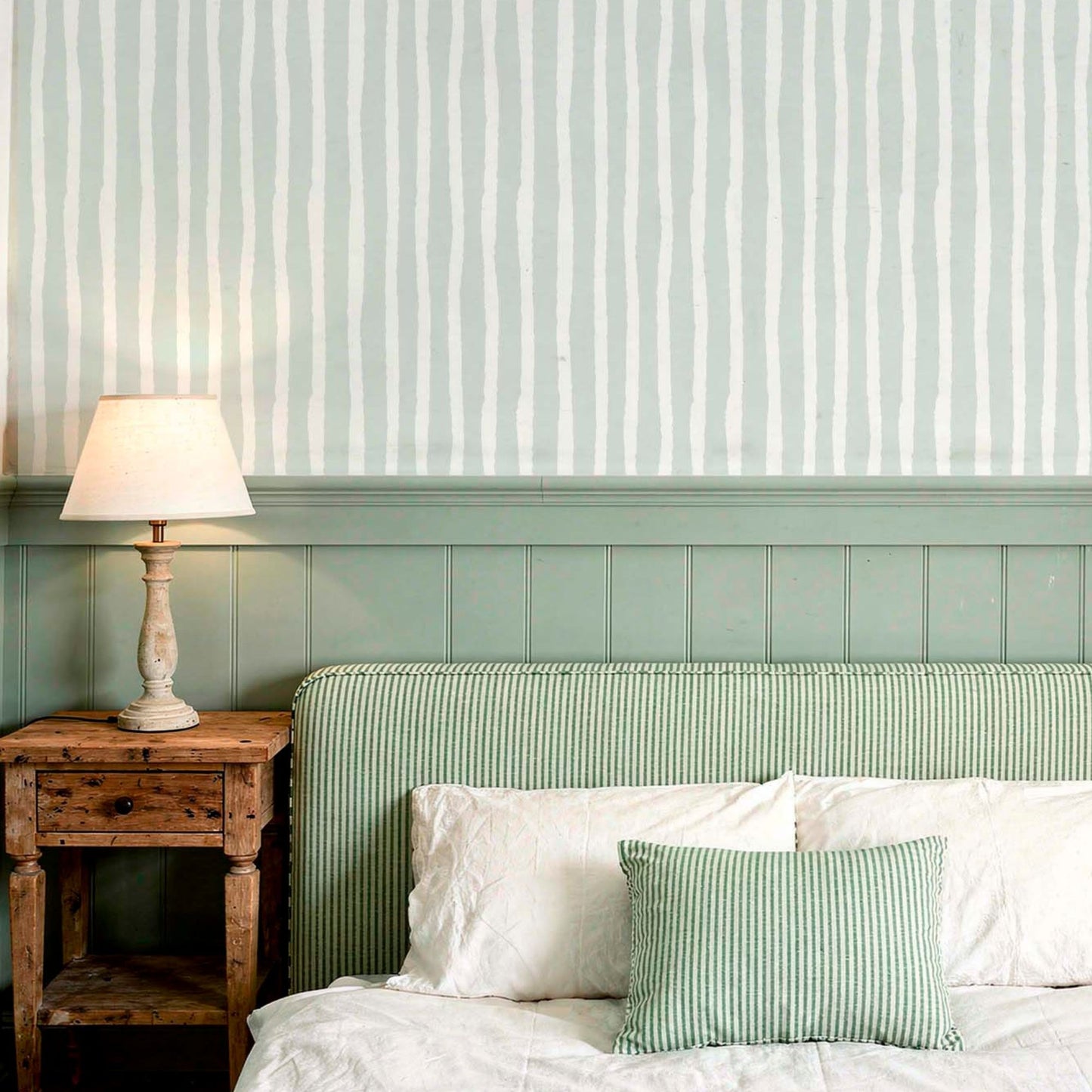 cat-616983200072 A cozy, rustic bedroom featuring a wall with sage green wood paneling and wavy, hand-drawn vertical striped wallpaper above it. A weathered wood nightstand holds a small glowing lamp next to a bed with a striped green headboard and matching decorative pillows, creating a calm and natural atmosphere.