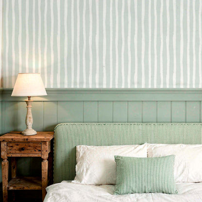 cat-616983200072 A cozy, rustic bedroom featuring a wall with sage green wood paneling and wavy, hand-drawn vertical striped wallpaper above it. A weathered wood nightstand holds a small glowing lamp next to a bed with a striped green headboard and matching decorative pillows, creating a calm and natural atmosphere.