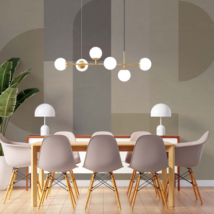 cat-610512830792 A modern dining room featuring a wooden table surrounded by six light grey Eames-style chairs with wooden legs. A brass linear chandelier with six white globe bulbs hangs above the table. The background features a large-scale geometric wall mural in muted earth tones of sage, taupe, and beige, with two white mushroom-style table lamps sitting on a side console