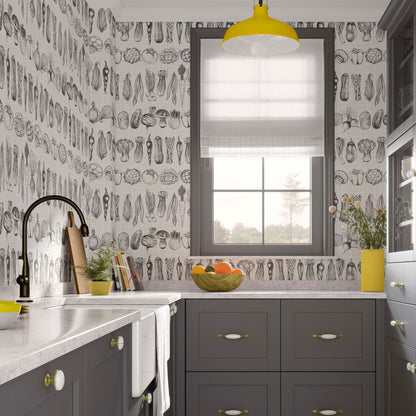 Modern kitchen with charcoal grey cabinets, white marble countertops, and wallpaper featuring black and white hand-drawn vegetable sketches. A bright yellow pendant light hangs above a window with a grey frame.