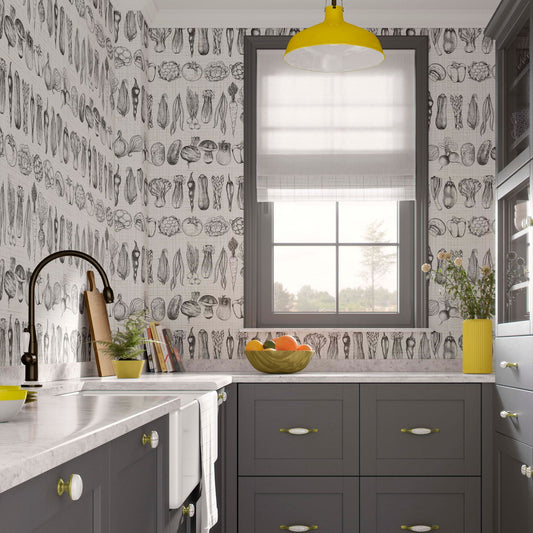 Modern kitchen with charcoal grey cabinets, white marble countertops, and wallpaper featuring black and white hand-drawn vegetable sketches. A bright yellow pendant light hangs above a window with a grey frame.