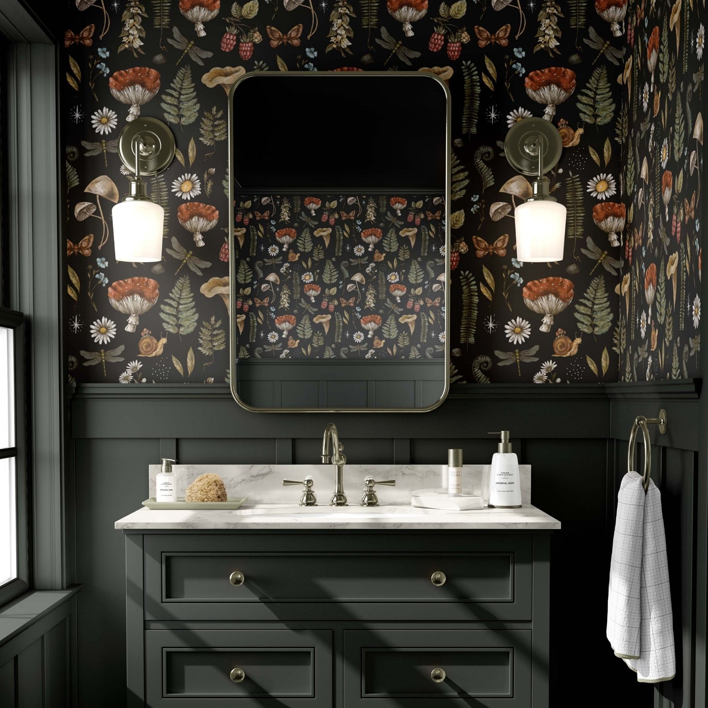 cat-610804236616 A moody and sophisticated bathroom featuring an enchanting forest wallpaper with detailed illustrations of mystical mushrooms, ferns, dragonflies, and butterflies on a deep black background. The dark botanical pattern creates a scholarly, dark cottagecore atmosphere above charcoal-grey wainscoting and a matching vanity with a marble countertop. Two antique brass sconces flank a rounded rectangular mirror, casting a warm glow in this atmospheric space.