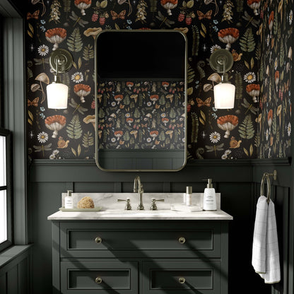 cat-610804236616 A moody and sophisticated bathroom featuring an enchanting forest wallpaper with detailed illustrations of mystical mushrooms, ferns, dragonflies, and butterflies on a deep black background. The dark botanical pattern creates a scholarly, dark cottagecore atmosphere above charcoal-grey wainscoting and a matching vanity with a marble countertop. Two antique brass sconces flank a rounded rectangular mirror, casting a warm glow in this atmospheric space.