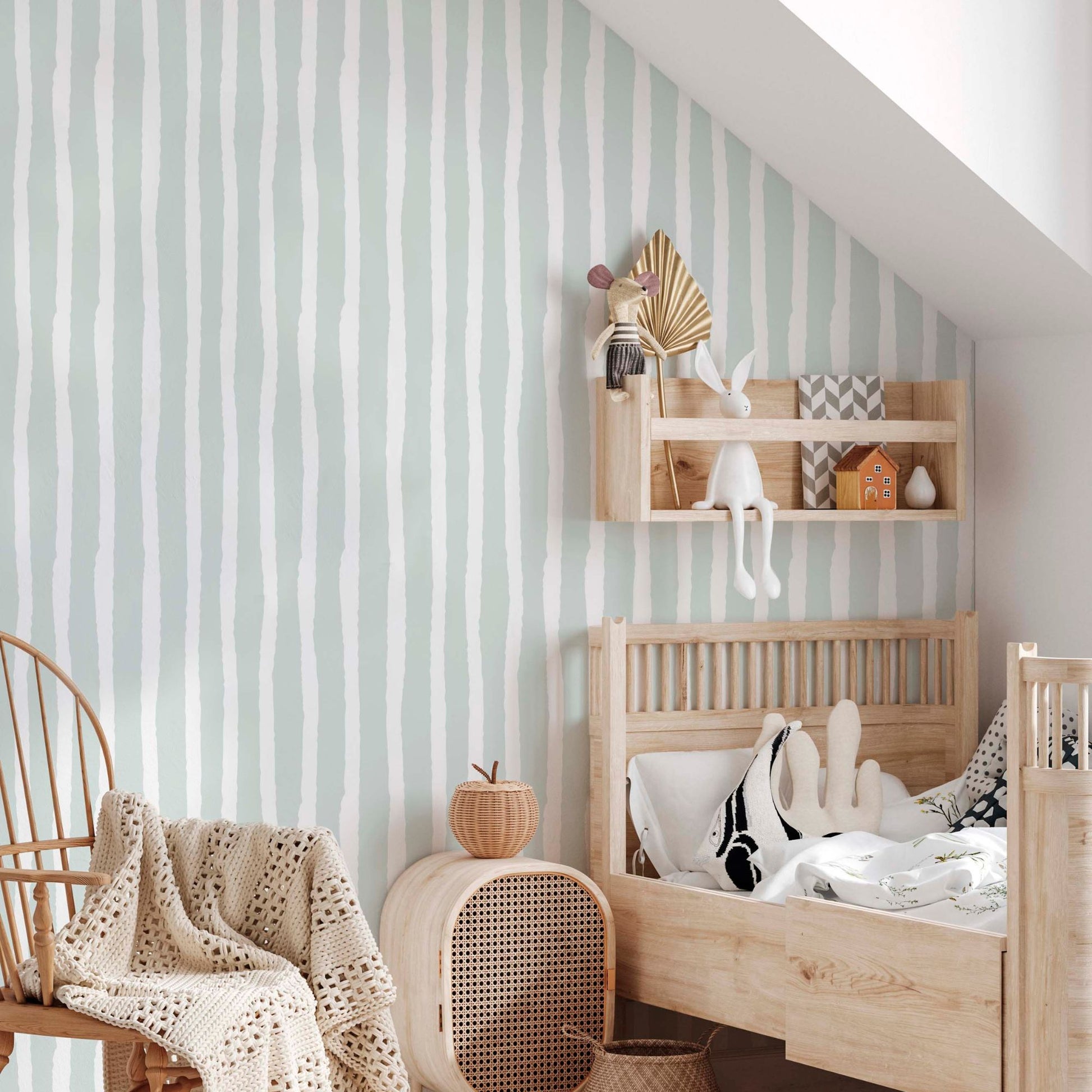 cat-615198294344 A bright, Scandinavian-style children's bedroom featuring mint green and white wavy-striped wallpaper. The room is decorated with light natural wood furniture, including a toddler bed, a wall-mounted shelf with toys, and a rattan chair with a knit throw blanket, creating a peaceful and warm nursery aesthetic.