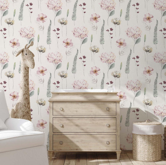 A cozy bedroom showing a natural wood dresser , a wicker laundry basquet and a white arm chair. The pink floral wallpaper adds a touch of nature with its delicate flowers and greens.