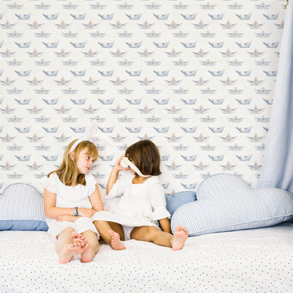 Two young girls sitting on a bed in front of a white wall decorated with paper boat pattern wallpaper.