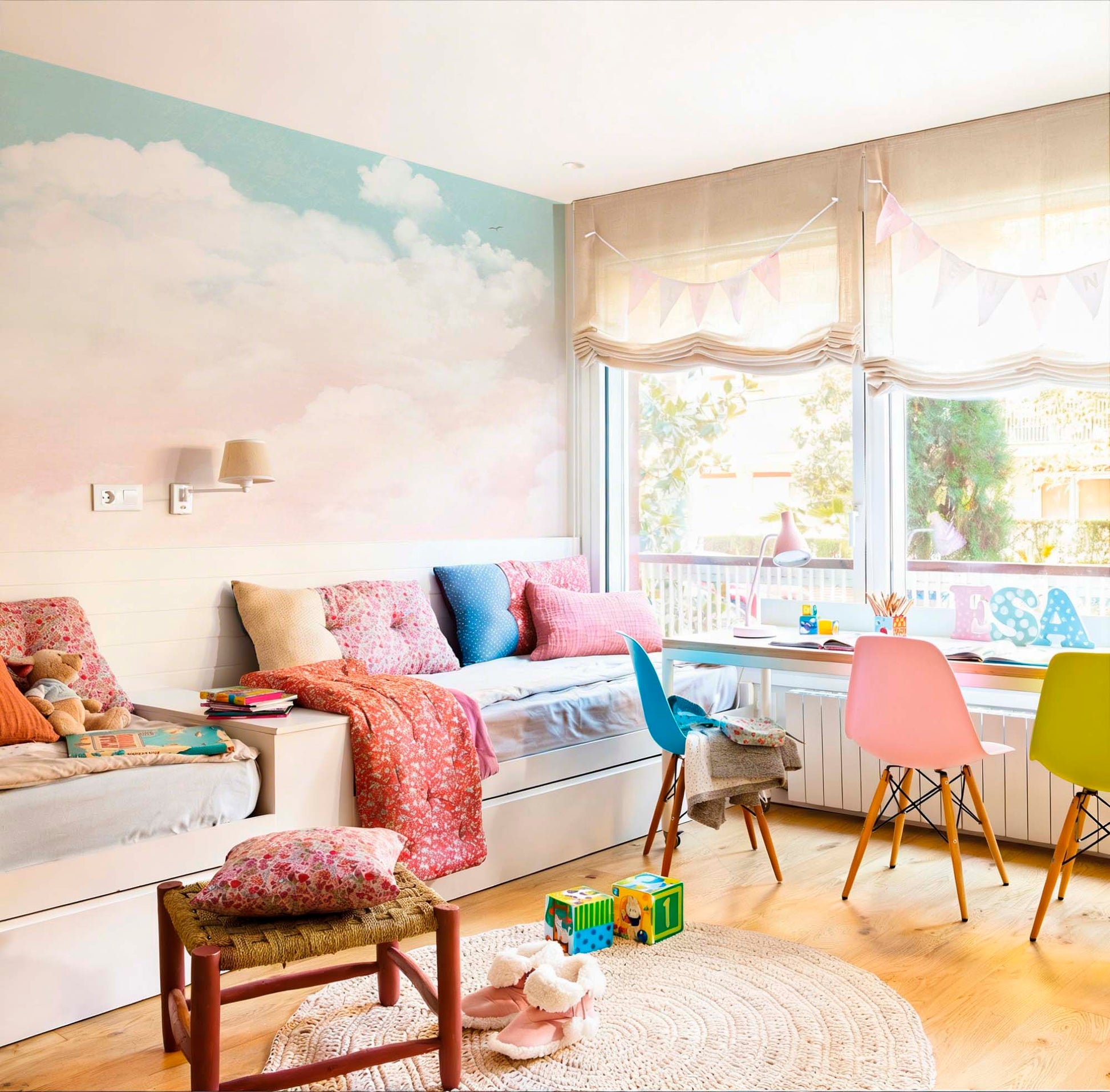 Pastel sky and clouds wallpaper mural in a kids' bedroom with white daybed, colorful chairs and jute rug