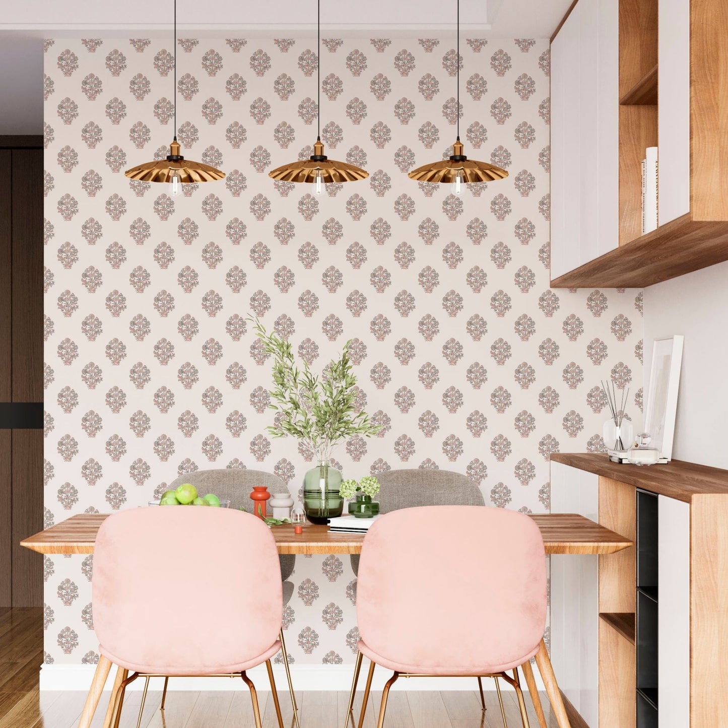 cat- 659356549448 A contemporary and stylish dining area featuring a pink floral block print wallpaper with symmetrical buta motifs. Two plush velvet blush pink chairs with slender gold legs are tucked under a warm wood dining table. Three matching fluted gold pendant lights hang above, beautifully complementing the botanical accent wall and the custom white and wood shelving unit, creating a warm and inviting space.