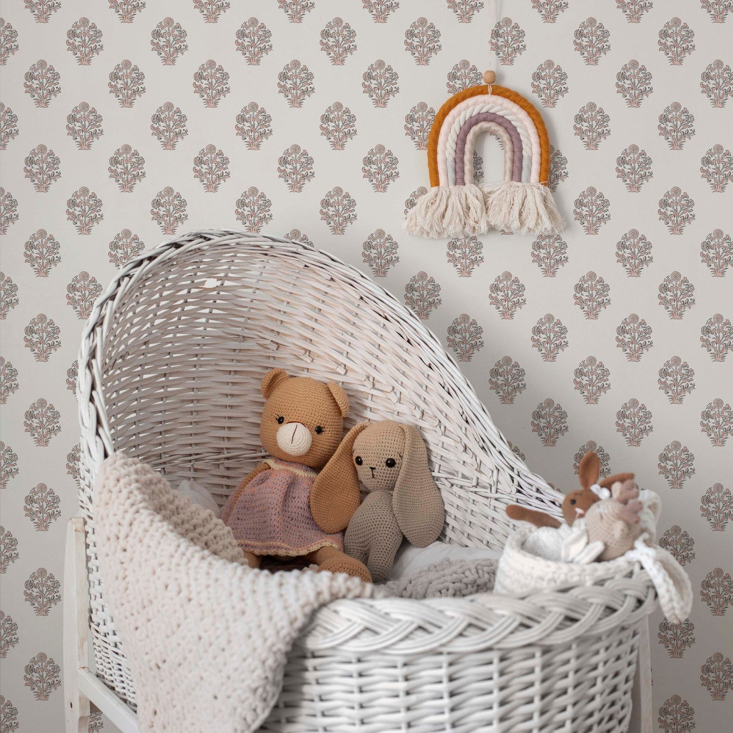 A cozy, neutral-toned nursery corner featuring a vintage-style floral block print wallpaper with soft pink and grey buta motifs. In the foreground, a white wicker bassinet is filled with a knitted cream blanket and two plush animal toys—a bear in a pink dress and a grey bunny. A colorful macramé rainbow wall hanging adds a whimsical touch to the serene, bohemian-inspired space.