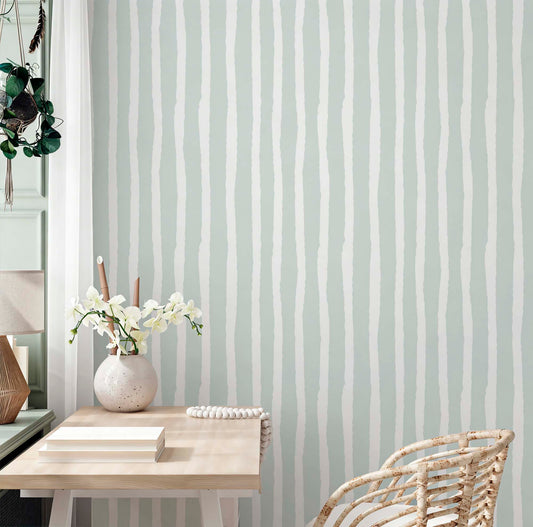 A serene interior scene featuring a wall covered in sage green and white wallpaper with hand-drawn, wavy vertical stripes. A light wood desk with books, a white ceramic vase of orchids, and a rattan chair sit in front of the wall, creating a modern boho aesthetic.
