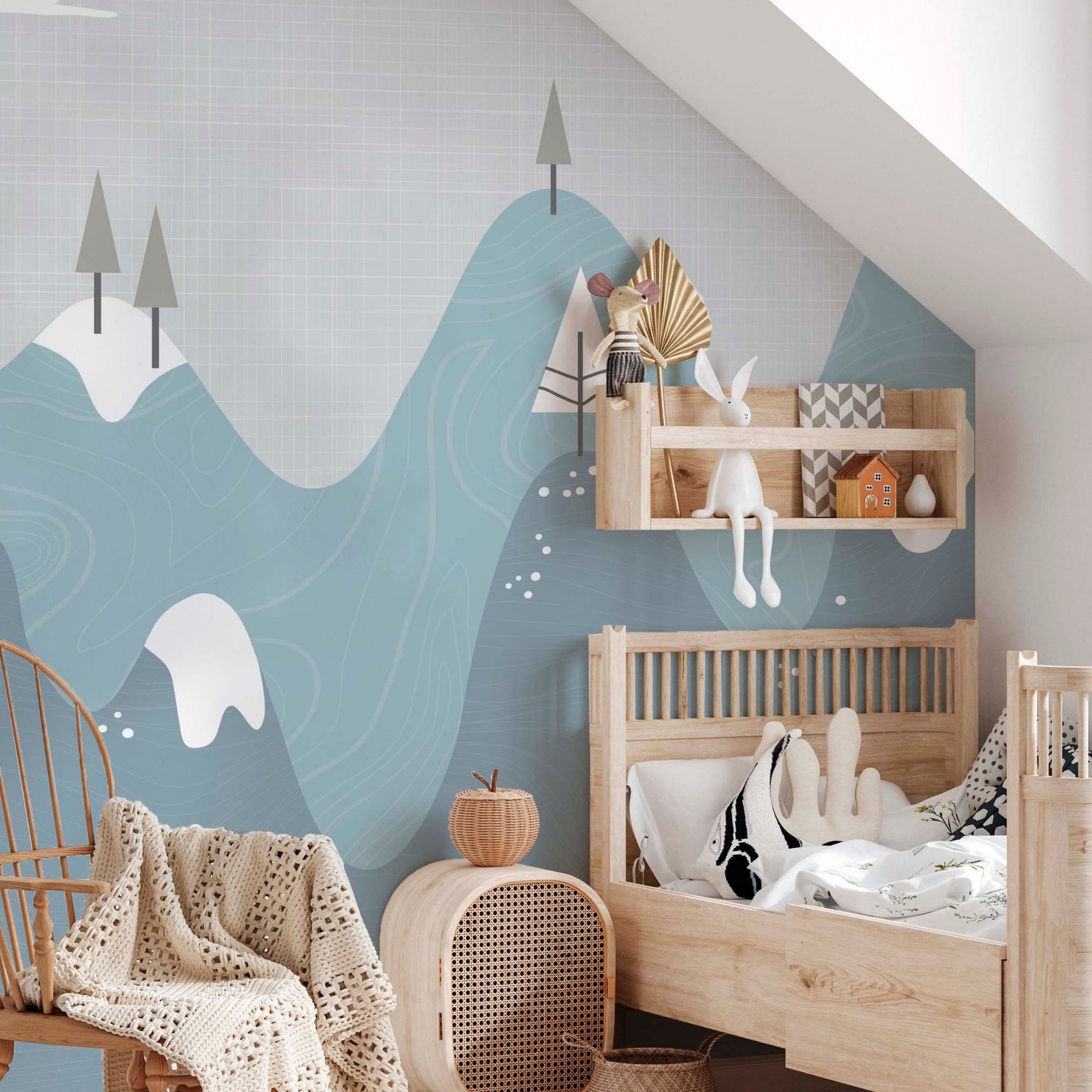 Snowy mountains nursery mural wallpaper in blue and grey tones in a nursery with wooden crib, rattan chair and natural wood shelf
