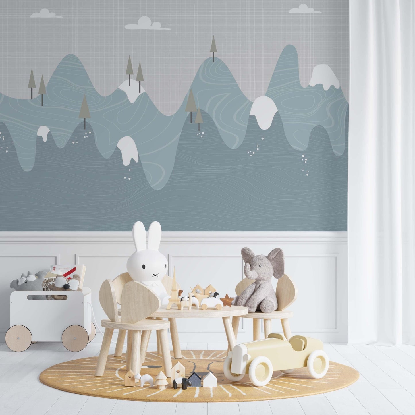 Snowy mountains nursery mural wallpaper in blue and grey tones in a playroom with white furniture, wooden chairs and soft toys
