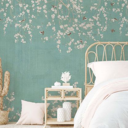 Boho-style bedroom featuring a teal wallpaper mural with white blossom branches and birds, a rattan headboard, and a light-wood nightstand with decorative coral.