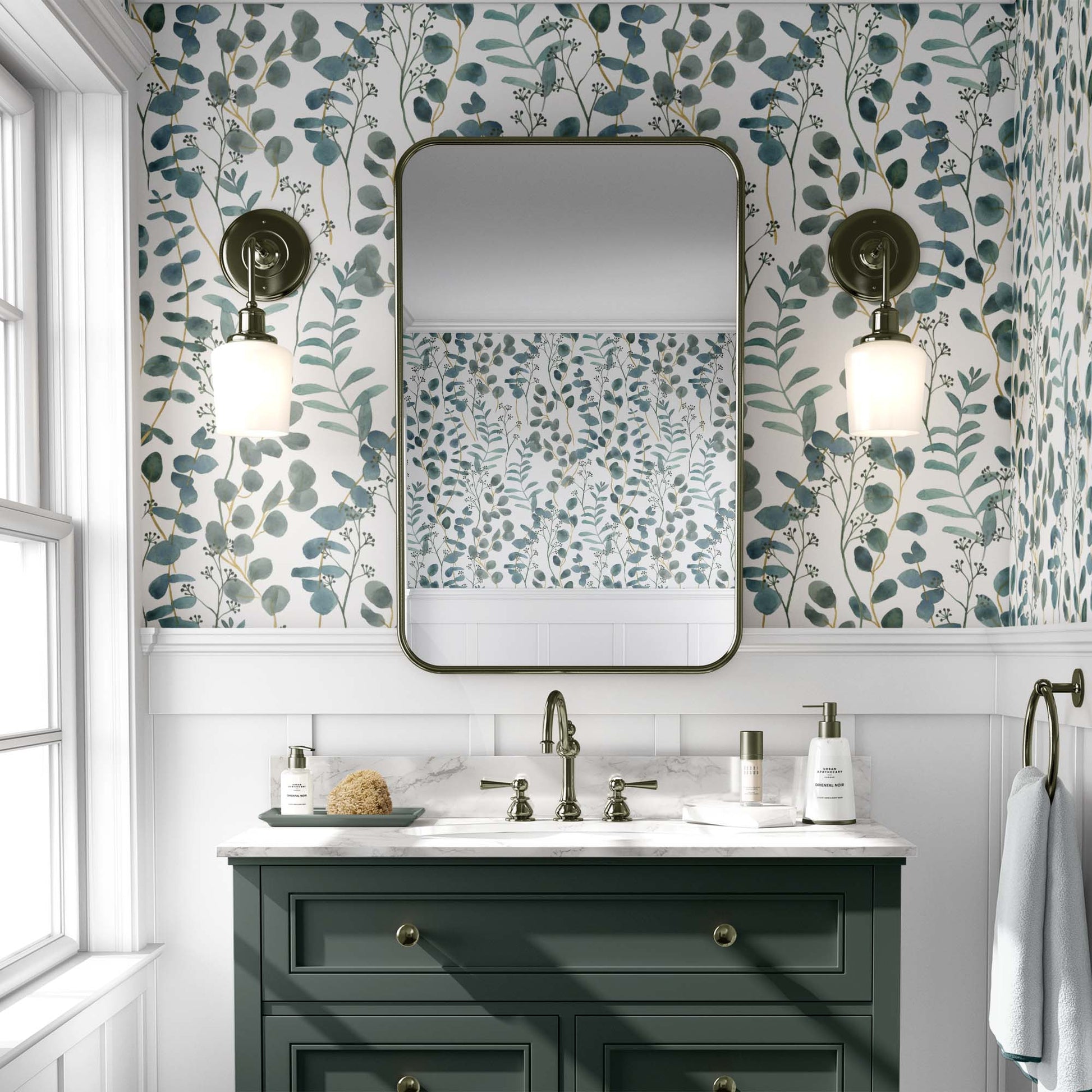 A bright bathroom featuring teal botanical wallpaper with watercolor eucalyptus leaves and branches. The floral pattern is paired with a dark green vanity, a white marble countertop, and a large rectangular mirror with rounded corners. Two antique-style brass wall sconces flank the mirror, reflecting the wallpaper's airy design on the opposite wall.