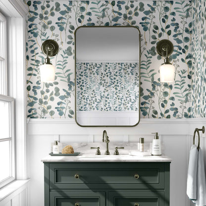 A bright bathroom featuring teal botanical wallpaper with watercolor eucalyptus leaves and branches. The floral pattern is paired with a dark green vanity, a white marble countertop, and a large rectangular mirror with rounded corners. Two antique-style brass wall sconces flank the mirror, reflecting the wallpaper's airy design on the opposite wall.