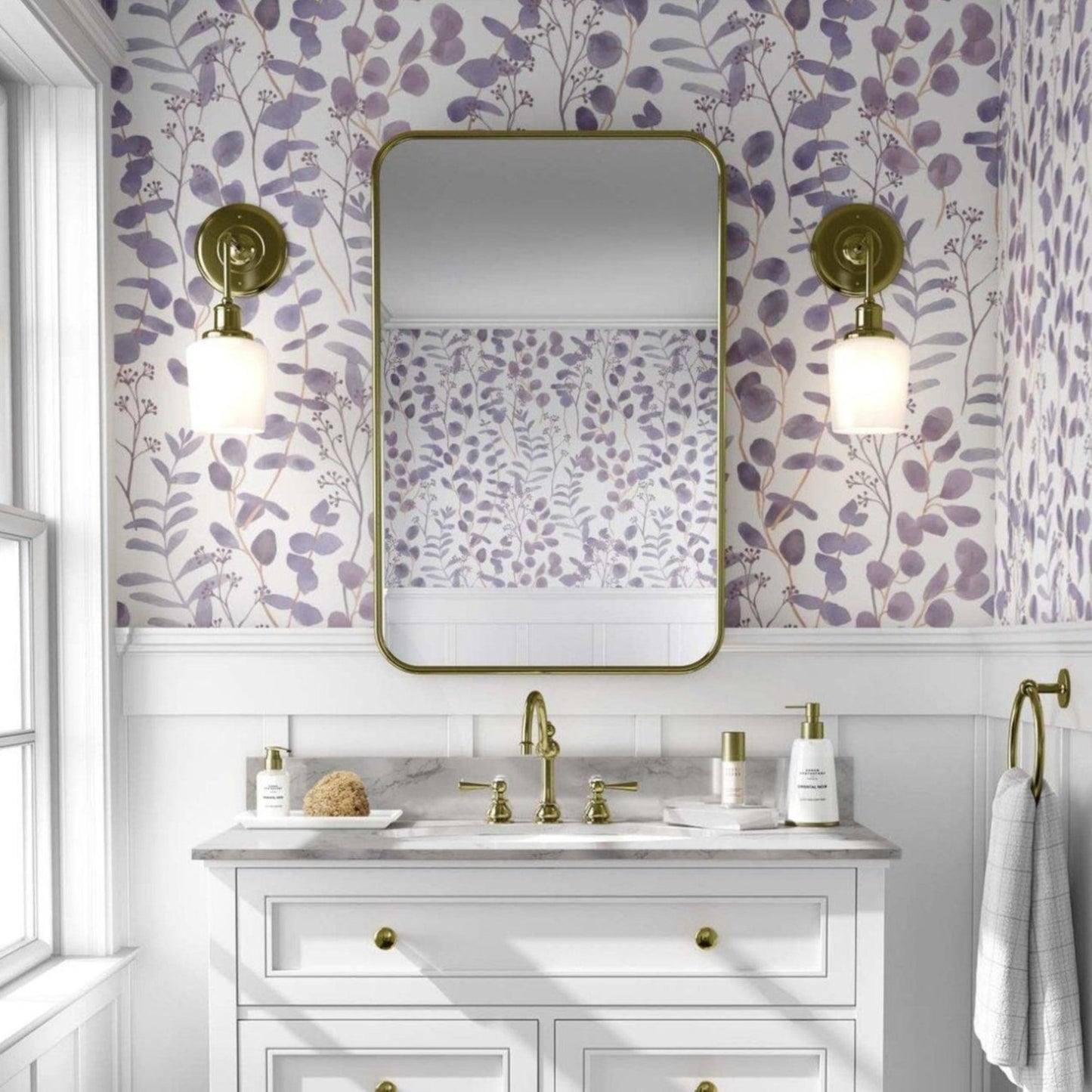 A traditional style bathroom featuring a white three-drawer vanity with a marble countertop. The upper wall is decorated with botanical lilac eucalyptus leaf wallpaper, complemented by a gold-framed rectangular mirror and two brass wall sconces with white glass globes.