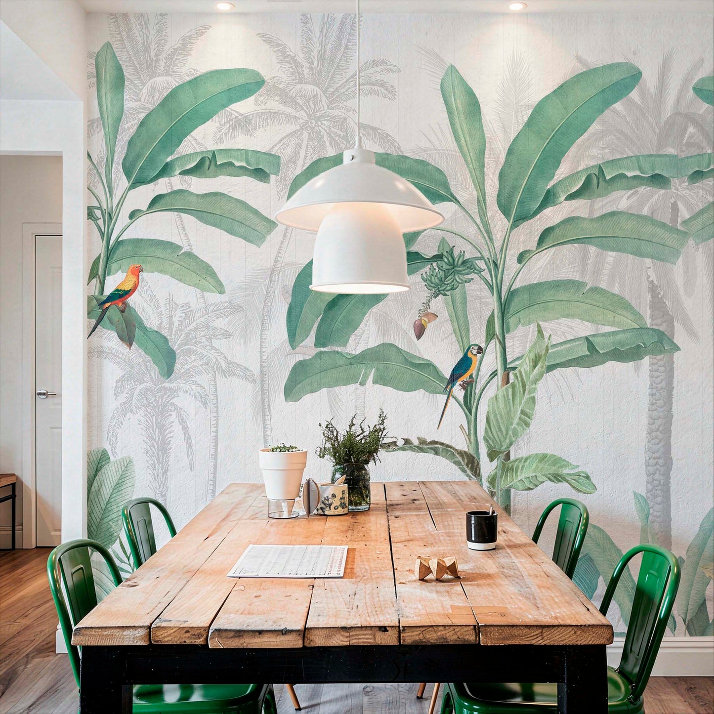 A large wall mural featuring vibrant green banana leaves, palm trees, and colorful parrots behind a rustic wooden dining table with green metal chairs.