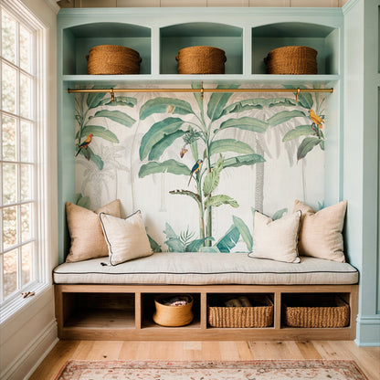 cat-610569683272 A custom mudroom built-in with light green cabinetry, a tropical banana leaf wallpaper mural backdrop, a cushioned bench with pillows, and wicker storage baskets