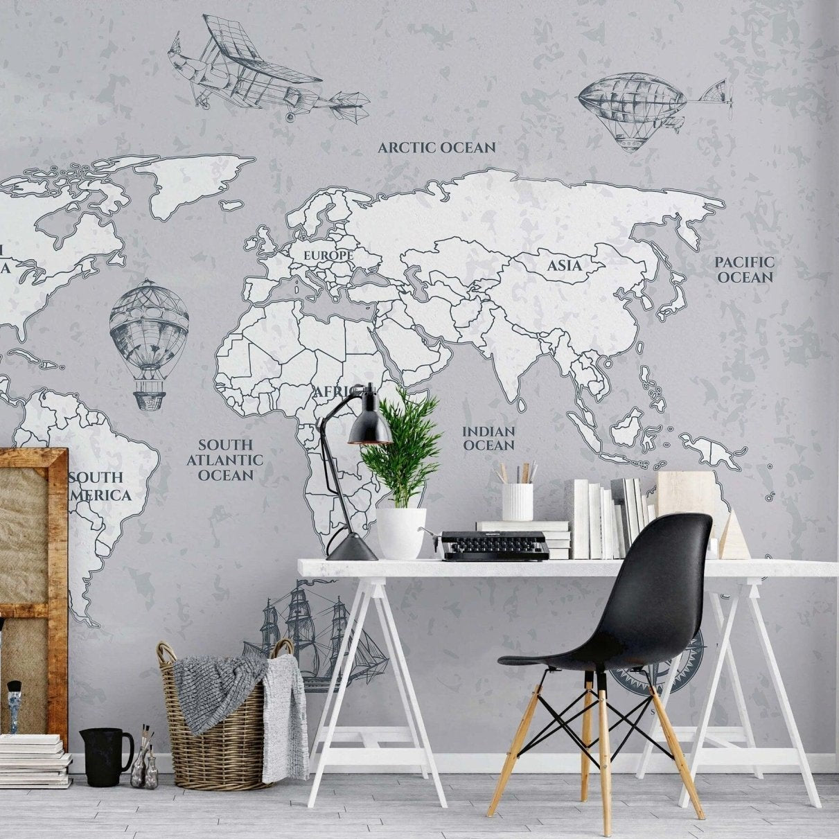 cat-660961526088 Monochrome vintage world map wallpaper mural installed on a home office wall behind a white trestle desk with a black Eames chair, typewriter, and leaning picture frames.