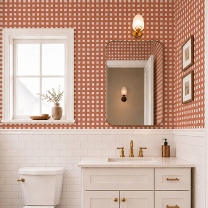 cat-610804236616 Terracotta check wallpaper in a bright bathroom with white subway tiles, a white vanity, brass fixtures and warm natural light.