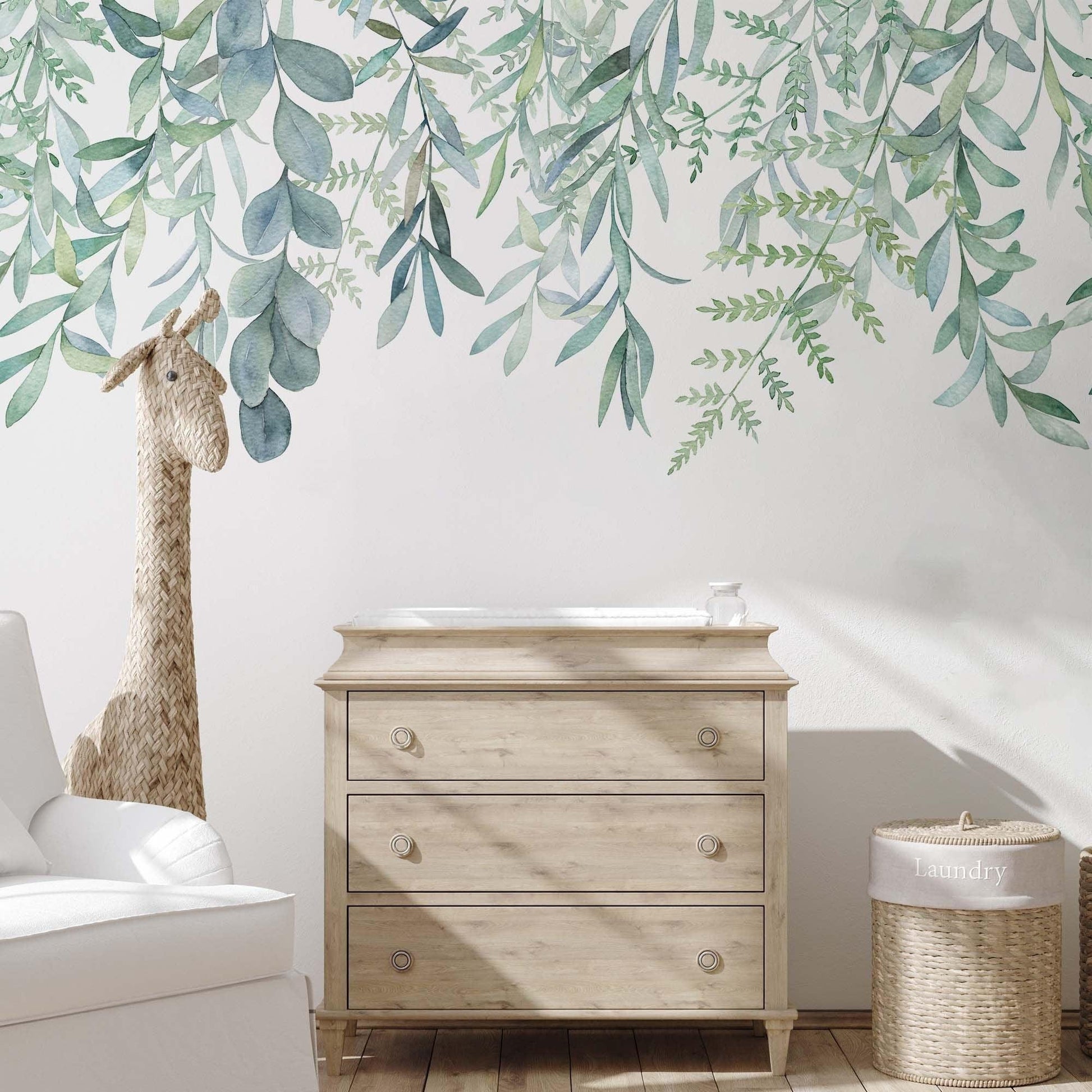 Hanging leaves wallpaper mural with soft green watercolour botanical leaves in a nursery with light wood dresser, rattan giraffe toy and wicker basket