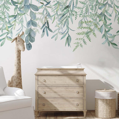Hanging leaves wallpaper mural with soft green watercolour botanical leaves in a nursery with light wood dresser, rattan giraffe toy and wicker basket